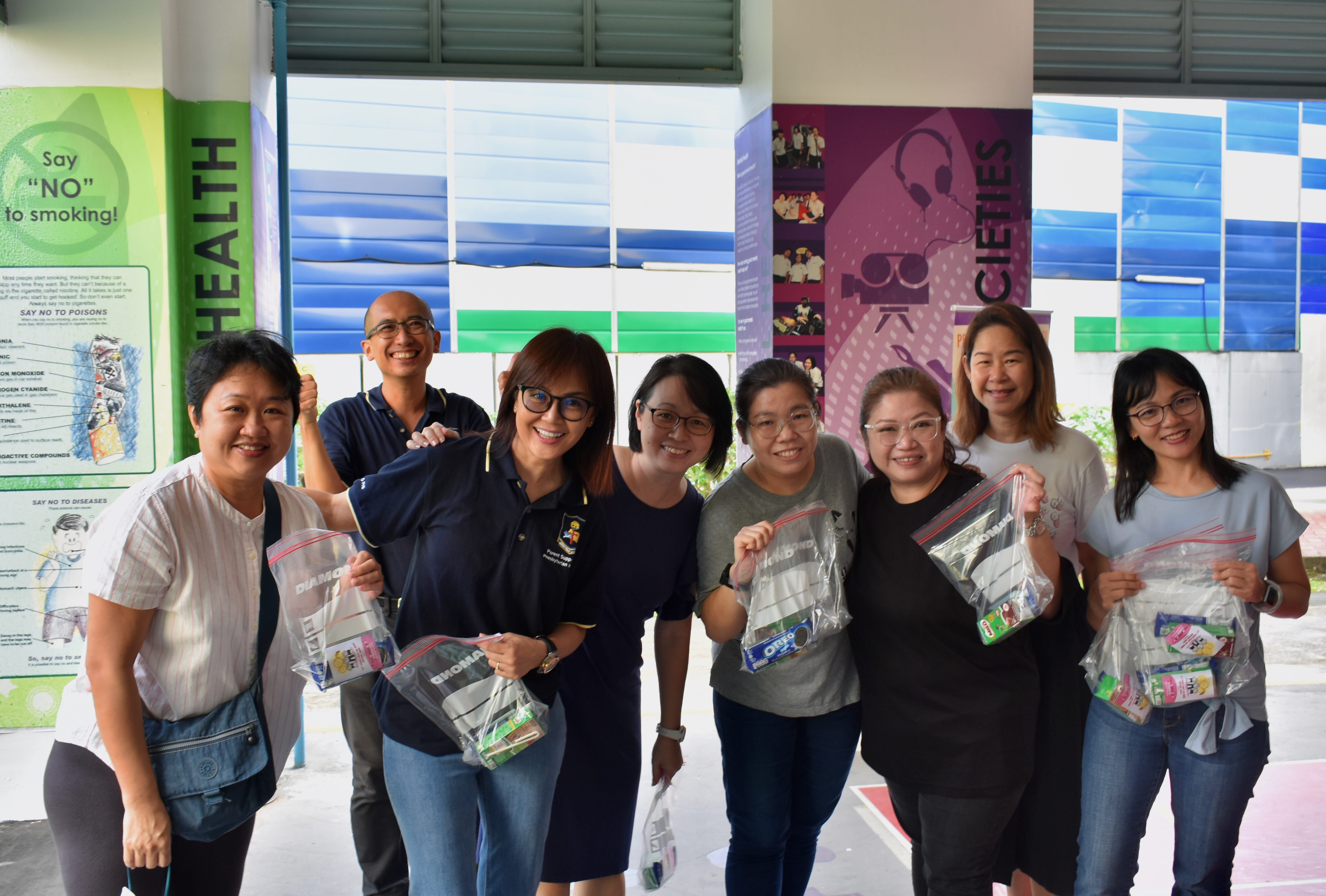 Eight smiling people stand together holding clear plastic bags with snacks, in front of a health awareness display.